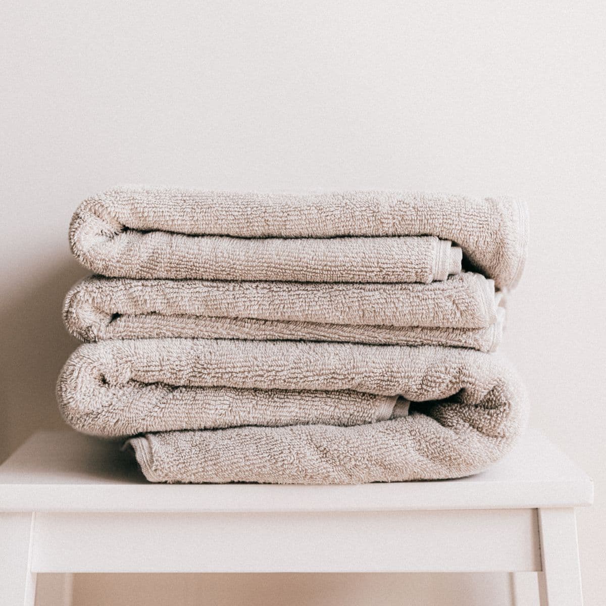 A folded stack of stone-neutral cotton bath towels on a white stool