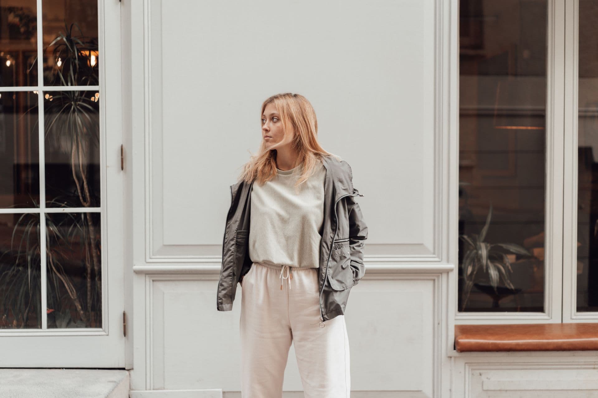 A model in a soft grey jacket and cream layers against a calm white facade